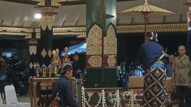 A man in traditional clothing sits near a decorated pillar at a ceremonial event with trophies on a table nearby.