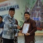Two men in batik shirts shake hands as one receives a certificate at a cultural ceremony, with others holding documents in the background and a decorative banner behind them.