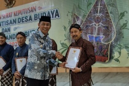 Two men in batik shirts shake hands as one receives a certificate at a cultural ceremony, with others holding documents in the background and a decorative banner behind them.