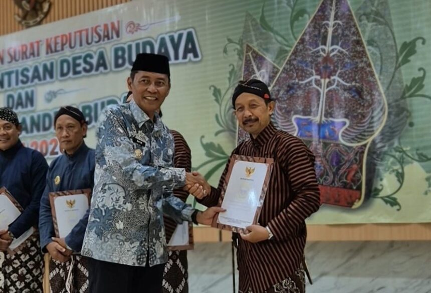 Two men in batik shirts shake hands as one receives a certificate at a cultural ceremony, with others holding documents in the background and a decorative banner behind them.