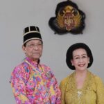 Elderly couple in traditional Malay attire posing for a formal portrait with a crest on the wall behind them.