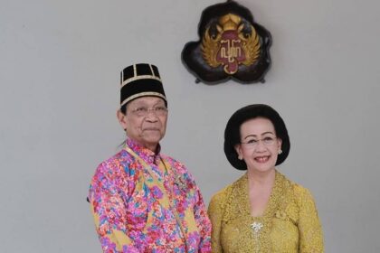 Elderly couple in traditional Malay attire posing for a formal portrait with a crest on the wall behind them.