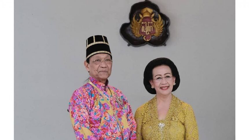 Elderly couple in traditional Malay attire posing for a formal portrait with a crest on the wall behind them.