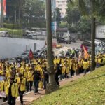 Large group of people wearing yellow jackets marching along a city street with colorful banners and flags.