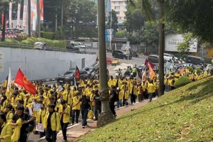 Large group of people wearing yellow jackets marching along a city street with colorful banners and flags.