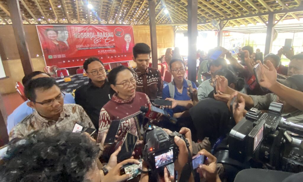 Crowded media scrum: reporters surround a speaker, holding microphones and smartphones indoors with a red banner in the background.