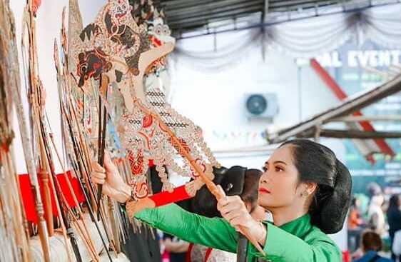 Woman in green adjusts ornate wooden shadow puppets on a display wall.