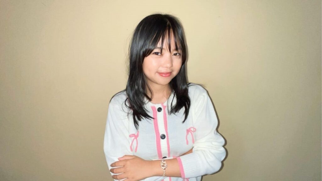 Smiling young woman with shoulder-length black hair, wearing a white cardigan with pink trim, standing against a beige wall with arms crossed.