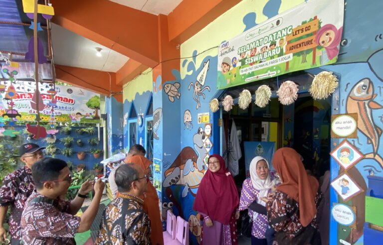 Colorful indoor entrance to a kids' activity area with banners and decorative hanging ornaments, people in batik clothing waiting in line.