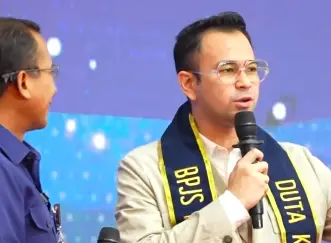 Man in a beige suit speaks into a microphone on a blue-stage backdrop, wearing a black-and-yellow scarf; another man in blue stands nearby during a talk.