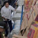 Three men inspect stacked cardboard boxes and white sacks inside a cargo truck in a loading area (supply shipment).