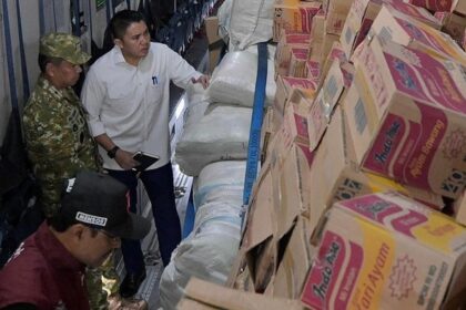 Three men inspect stacked cardboard boxes and white sacks inside a cargo truck in a loading area (supply shipment).
