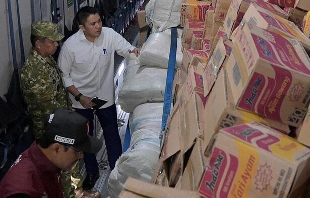 Three men inspect stacked cardboard boxes and white sacks inside a cargo truck in a loading area (supply shipment).