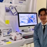 Man in a blue blazer stands beside a desk with an iMac showing a 'Catalogue' screen at a booth or exhibition.",