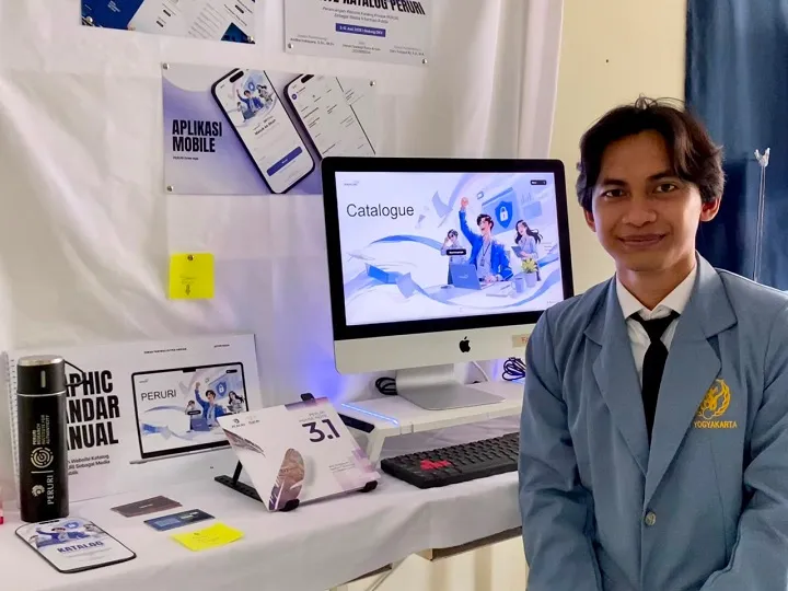 yyuui - Mabur.co Man in a blue blazer stands beside a desk with an iMac showing a 'Catalogue' screen at a booth or exhibition.",
