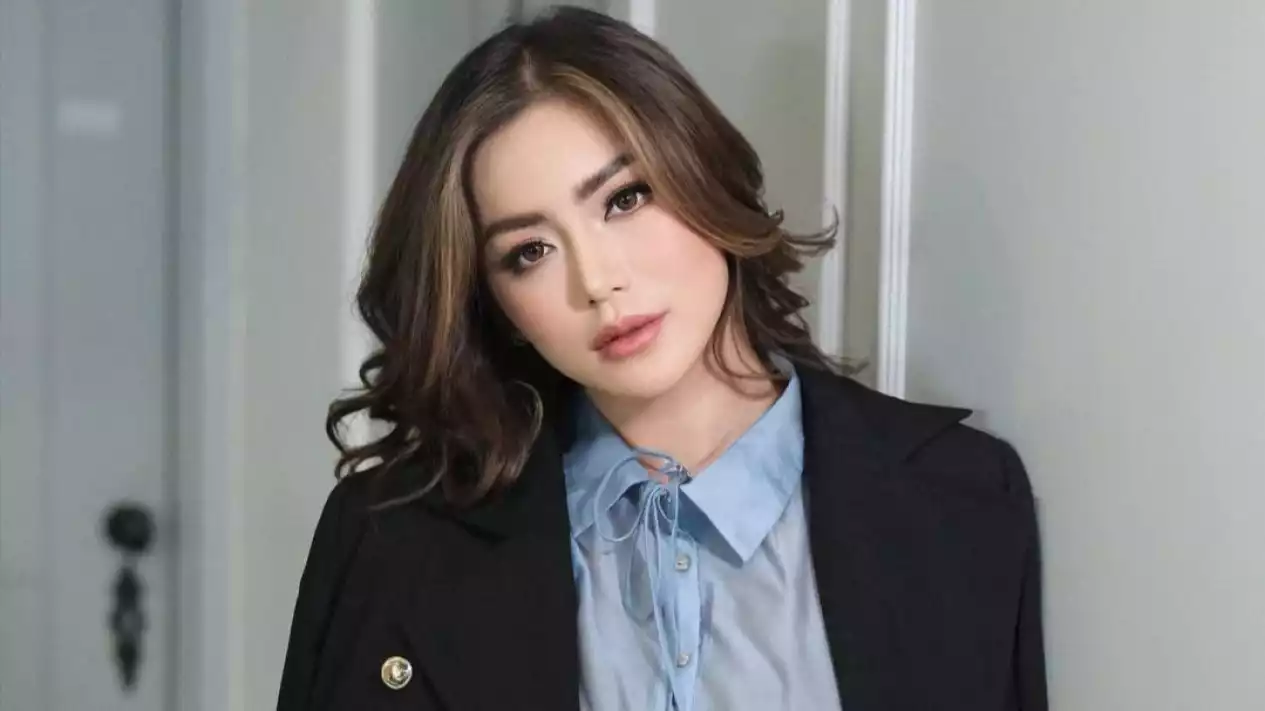 Portrait of a woman with shoulder-length wavy hair, wearing a light blue blouse and black blazer in an indoor setting.