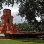 Red brick ancient tower ruin on a raised platform in a palm-lined courtyard garden