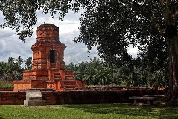 Red brick ancient tower ruin on a raised platform in a palm-lined courtyard garden