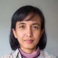 zaA - Mabur.co Portrait of a woman with short black hair, wearing a pink turtleneck and plaid shirt, looking at the camera.