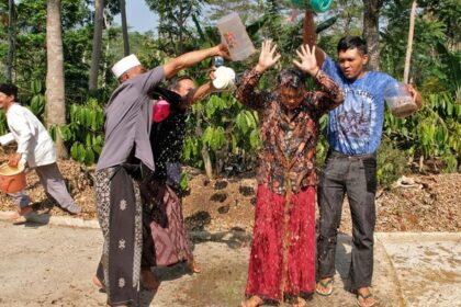 Group of people splash water from plastic buckets on a woman in a red skirt during a water festival in a tropical garden setting.
