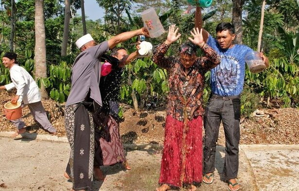 Group of people splash water from plastic buckets on a woman in a red skirt during a water festival in a tropical garden setting.