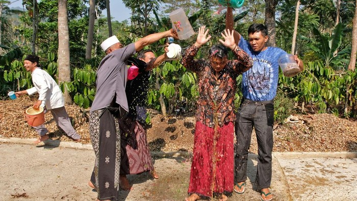 Group of people splash water from plastic buckets on a woman in a red skirt during a water festival in a tropical garden setting.