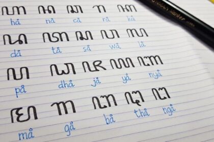 Notebook page with black script syllables and blue pronunciation guides, laid out in rows for writing practice