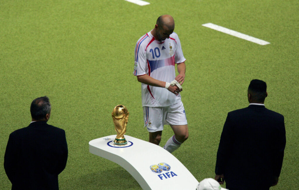 zidane-trophy-2006-scaled - Mabur.co Footballer wearing a white France jersey with number 10 stands on a podium beside the FIFA World Cup trophy on a green pitch, with suited officials nearby