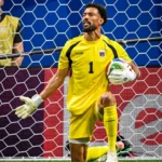Soccer goalkeeper in a yellow kit kneels by the goal, holding a ball under his right arm with gloves on.