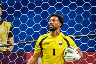 Soccer goalkeeper in a yellow kit kneels by the goal, holding a ball under his right arm with gloves on.