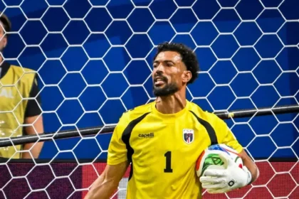Soccer goalkeeper in a yellow kit kneels by the goal, holding a ball under his right arm with gloves on.